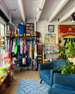 Interior of Amity's Maine Vintage in Portland, Maine colorful vintage clothing on a rack, retro dishware, housewares, handmade goods filling shelves, blue mid-century armchair and large retro art print.